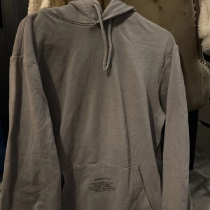 Grå hoodie från H&M - Säljer en stilren grå hoodie från H&M med tryck på framsidan och baksidan. Tröjan har en klassisk design med luva, samt en stor ficka framtill. Perfekt för en avslappnad look.