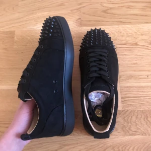 Christian Louboutin skor storlek 40 - Snygga svarta sneakers från Christian Louboutin med nitar på tån och en ikonisk röd sula. Skorna har snörning och är tillverkade i mocka.