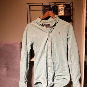 Grön/ljus blå skjorta från Ralph Lauren - Snygg skjorta från Ralph Lauren i slim fit. Tillverkad i stretchigt oxfordtyg med klassiska knappar och broderad logotyp på bröstet. Perfekt för en stilren look! Säljer pågrund ut av att den börjar bli liten. Pris kan diskuteras 