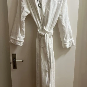 Vit badrock i velour - Mysig vit badrock i mjuk velour från märket C. Den har långa ärmar och ett praktiskt knytbälte i midjan för en bekväm passform. Perfekt för att svepa in sig efter duschen eller en avslappnad morgon. Kostar 1000kr på Mio 
