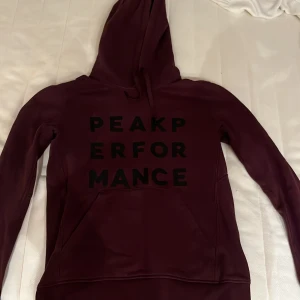 Vinröd hoodie från Peak Performance - Säljer en snygg vinröd hoodie från Peak Performance med svart text på framsidan. Den har en klassisk känguruficka och justerbar huva med dragsko. Perfekt för en avslappnad stil.