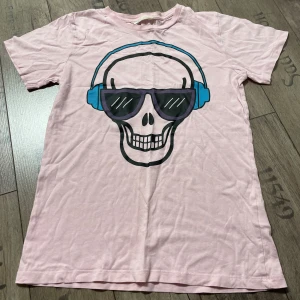 Rosa t-shirt med döskalletryck från H&M - Cool rosa t-shirt från H&M med ett stort tryck av en döskalle som bär hörlurar och solglasögon. Perfekt för en avslappnad stil med en twist. Tillverkad i mjukt material för bekväm passform.