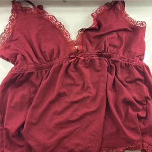 Vinröd topp med spetsdetaljer från Nelly - Säljer en vinröd topp från Nelly med vackra spetsdetaljer längs kanterna. Toppen har en söt rosett framtill. Kan knytas där bak om det behövs men kommer utan knyten axelband. Är lite knottrig framifrån men syns knappast. Använd fåtal gånger och är en väldigt fin topp till många tillfällen. Kommer tvättats och strykas innan den skickas så den kommer i ett fint skick. Köptes för 250kr och säljs för 150kr plus frakt.