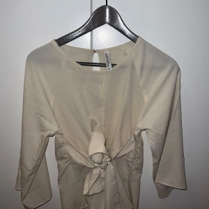 Beige blus med knyt - Snygg beige blus med knyt framtill. Blusen har trekvartsärmar och en rund halsringning. Perfekt för en stilren look.