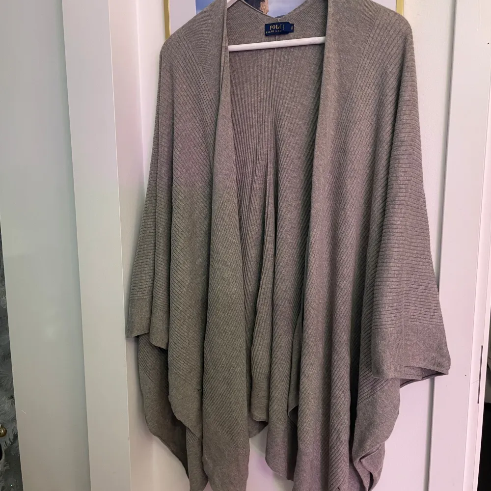 Säljer en stilren grå poncho från Polo Ralph Lauren. Den är ribbad och har en öppen design utan knappar eller dragkedja. Ponchon har några defekter som lätt kan sys och som inte syns när denna är på en!. Asusteet.