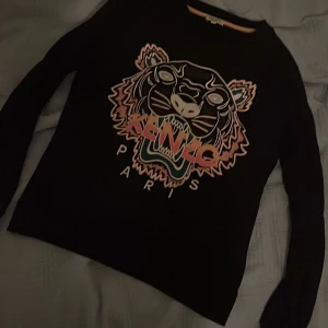 Svart tröja med tigertryck från Kenzo - Snygg svart tröja från Kenzo med ett coolt tigertryck i flera färger och texten 'Paris'. Perfekt för att ge din outfit en trendig touch. Tröjan har långa ärmar och en normal passform.