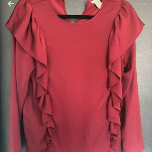 Röd tröja med volangdetaljer - Snygg röd tröja med volangdetaljer längs sidorna. Perfekt för att ge en extra touch till din outfit. Tröjan har långa ärmar och en rund halsringning.