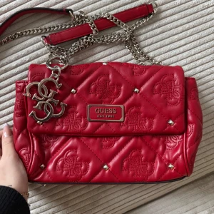 Röd quiltad axelväska från Guess - Snygg röd axelväska från Guess med quiltat mönster och silverdetaljer. Väskan har en kedjerem och ett stort fack med dragkedja inuti. Perfekt för att ge din outfit en färgklick! Aldrig använd, så den är helt ny utan prislapp. Köpte för 1200 kr på zalando men säljer för 399 kr.