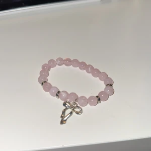 Rosa armband med rosettberlock - Snyggt rosa armband med pärlor och en söt rosettberlock i silverfärg. Perfekt accessoar för att ge en feminin touch till din outfit.