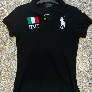 Svart pikétröja från Ralph Lauren - Snygg svart pikétröja från Ralph Lauren med italiensk flagga och texten 'ITALY' på bröstet. Stort vitt nummer 3 på ryggen och ärmen. Perfekt för en sportig look!