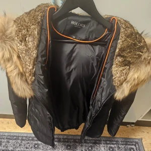 Svart jacka med pälsdetaljer från Hollies - Snygg svart jacka från Hollies med lyxig pälsdetalj på huvan. Perfekt för kyliga dagar med sin stilrena design och varma material. Jackan har långa ärmar och en praktisk dragkedja framtill.