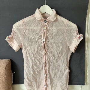 Ljusrosa kortärmad skjorta med unika knappar - Säljer en ljusrosa kortärmad skjorta med unika, mönstrade knappar. Skjortan har en klassisk krage och är tillverkad i ett lätt material, perfekt för varmare dagar. Detaljerade knappar ger en extra touch till den annars enkla designen.