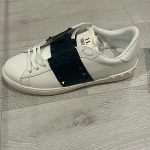 Valentino Garavani sneakers - Snygga vita Valentino Garavani sneakers med en svart detalj över snörningen. Skorna har en stilren design med diskreta nitar på sulan och är perfekta för en trendig look.