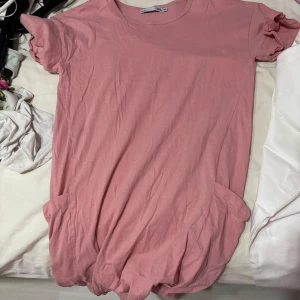 Rosa t-shirtklänning från 157 - Säljer en söt rosa t-shirtklänning från 157. Den har korta ärmar med volangdetaljer och en avslappnad passform. Perfekt för en bekväm och stilren look.
