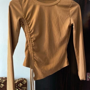 Brun ribbad topp med snörning - Säljer en snygg brun ribbad topp med lång ärm och cool snörning på sidan. Perfekt för höst och vår, den har en tight passform som framhäver figuren. Toppen är i ett mjukt och stretchigt material, vilket gör den superbekväm att ha på sig. Passar både till vardags och fest!