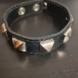 Svart armband med nitar - Säljer ett coolt svart armband med silverfärgade nitar. Armbandet har en justerbar knäppning med tryckknappar och är perfekt för att ge din outfit en edgy touch. Passar både till vardags och fest!