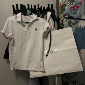 Vit pikétröja från Moncler - Säljer en snygg vit pikétröja från Moncler. Den har en klassisk krage med röd och blå randdetalj och korta ärmar. Moncler-loggan är broderad på bröstet. Perfekt för en stilren och avslappnad look!