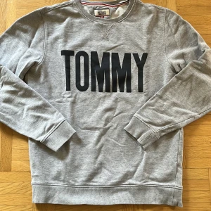 Grå sweatshirt från Tommy Hilfiger - Säljer en snygg grå sweatshirt från Tommy Hilfiger Denim. Den har ett stort svart tryck med texten 'TOMMY' på framsidan. Tröjan är i storlek M och har långa ärmar med ribbade muddar. Perfekt för en casual look eller en chill dag hemma. Superbekväm och stilren!