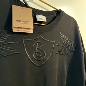 Säljer en snygg svart t-shirt från Burberry med ett coolt broderat emblem på bröstet. T-shirten har korta ärmar och är perfekt för både vardag och fest. Den är i nyskick och redo att bäras av någon som älskar stil och kvalitet!