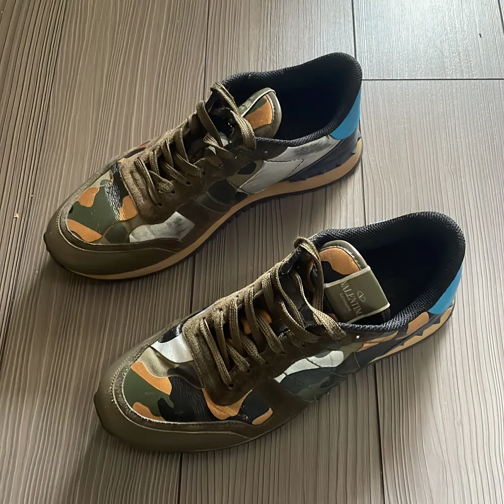 Säljer ett par snygga sneakers från Valentino i camouflage-mönster. Skorna har en cool mix av gröna, bruna och svarta nyanser med en pop av blått på hälen. De är i skinn och har snörning framtill. Perfekta för en casual look med en twist!. Skor.