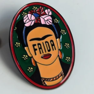 Färgglad Frida Kahlo brosch - Brosch med Frida Kahlo-motiv. Den är ovalformad och har en färgglad design med gröna, rosa, röda och svarta detaljer.NY