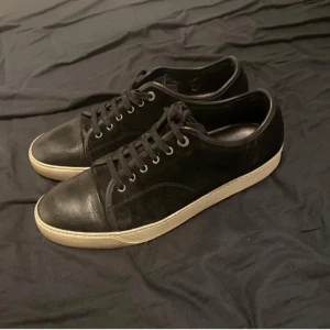 Svarta sneakers från Lanvin - Säljer ett par stilrena svarta sneakers från Lanvin. De har en snygg kombination av mocka och skinn med klassisk snörning. Sulan är i en kontrasterande beige färg som ger en cool look. Perfekta för både vardag och fest!