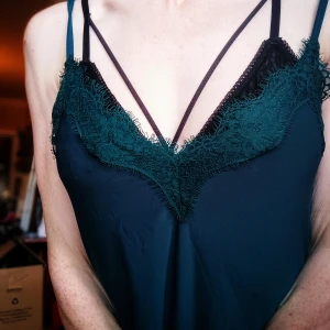 Mörk grönt  linne med spetsdetaljer - Snyggt mörk grönt linne med smala axelband och vacker spetsdetalj vid urringningen. Perfekt för en kväll ute eller en fest. Materialet är mjukt och följsamt, vilket gör det bekvämt att bära hela dagen. Passar bra till både jeans och kjol för en stilren look.