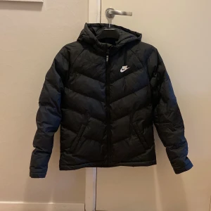 Svart dunjacka från Nike - Säljer en svart dunjacka från Nike i nyskick. Den har en snygg pufferdesign med dragkedja och huva, perfekt för kalla dagar. Jackan är långärmad och har en normal passform. Superbra för både höst och vinter!