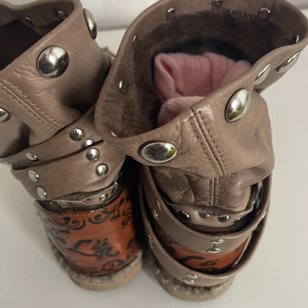 Säljer ett par unika beige boots i skinn med coola nitar och broderade detaljer på hälen. De har en avslappnad passform och är perfekta för höst och vår. Skorna har en dekorativ rem med spänne och är i välanvänt skick.. Skor.