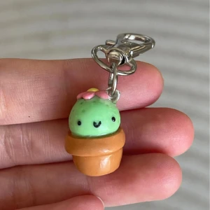 Nyckelring med kaktus i kruka - Supergullig nyckelring med en liten kaktus i en brun kruka. Kaktusen är ljusgrön med en söt rosa blomma på toppen och ett litet ansikte. Perfekt för att piffa upp dina nycklar eller väskan! Den är ungefär 4 cm lång inklusive metallklämman.