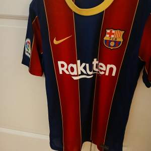 Säljer en FC Barcelona fotbollströja från Nike. Den är blå och röd med klubbens emblem på bröstet och Rakuten som sponsor. Tröjan har korta ärmar och La Liga-märke på sidan. Perfekt för fans som vill visa sitt stöd!