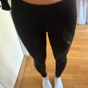 Svarta leggings från 2XU - Säljer ett par svarta leggings från 2XU i storlek dam XS. De är perfekta för träning med sitt tighta passform och snygga prickiga mönster längs benen. Materialet är syntetiskt och andas bra, vilket gör dem bekväma under intensiva pass. De är i nyskick och redo för nya äventyr!