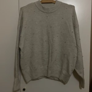  tröja med pärlor - Säljer en mysig grå/beige tröja från H&M i storlek S. Den är dekorerad med små pärlor över hela ytan för en extra touch. Tröjan har långa ärmar och en ribbad kant vid halsen. Observera att det finns ett litet hål på framsidan. 