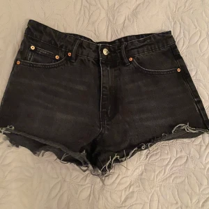 Svarta jeansshorts - Säljer ett par svarta jeansshorts i nyskick. De är högmidjade med en rå kant nertill för en cool look. Perfekta för sommaren eller en avslappnad dag ute. De har klassiska fem fickor och knappar i metall. Passar perfekt till en t-shirt eller linne!