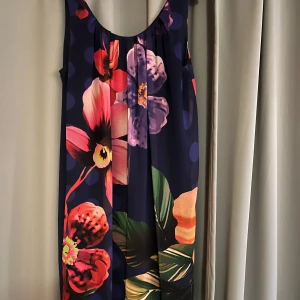 Blommig klänning från Desigual - Snygg blommig klänning från Desigual i mörkblått med stora färgglada blommor. Klänningen är ärmlös och har en lös passform, perfekt för en somrig look. Den har en rund halsringning och är tillverkad i ett mjukt material.