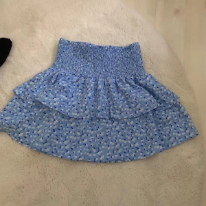 Blå blommig kjol från UNQ Young - Söt blå kortkjol från UNQ Young med ett blommigt mönster i vitt och blått. Kjolen har en elastisk midja och volangdetaljer som ger en lekfull look. Perfekt för varma dagar! Endast använd 2 gånger 