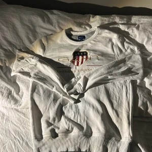 Vit sweatshirt från GANT - Säljer en vit sweatshirt från GANT med ett broderat emblem och texten 'American Sportswear' på framsidan. Tröjan har långa ärmar och ribbade muddar vid ärmslut och nederkant. Perfekt för en avslappnad stil. Den har en liten fläck vid ärmslut. Pris kan diskuteras.