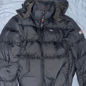 Svart dunjacka från Tommy Hilfiger - Säljer en svart dunjacka från Tommy Hilfiger med huva och dragkedja. Jackan har en klassisk pufferdesign och små logodetaljer på bröstet och ärmen. Perfekt för kalla dagar!