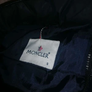 Svart dunväst från Moncler - Säljer en snygg svart dunväst från Moncler med glansig finish. Västen har en dragkedja framtill och Moncler-logga på bröstet. Perfekt för lager-på-lager under kyliga dagar. Kom med  bud ställ frågor om ni vill