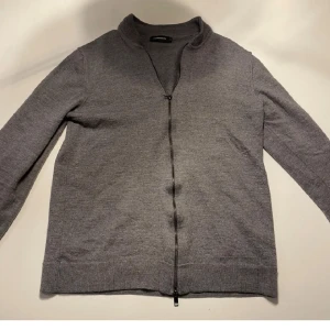 Grå kofta från J.Lindeberg - Snygg grå kofta från J.Lindeberg med dragkedja framtill. Perfekt för lager-på-lager och har en stilren design. Passar bra till både vardag och mer uppklädda tillfällen. Dragkedjans hände är av på den längst upp.