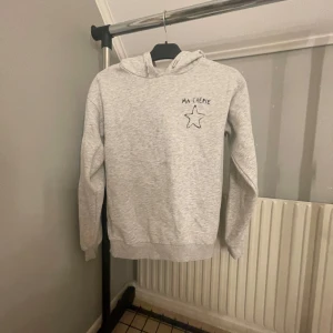 Grå hoodie med stjärndetalj - Säljer en grå hoodie med texten 'MA CHERIE' och en stjärna tryckt på både fram- och baksidan. Tröjan har en bekväm passform med långa ärmar och en klassisk huva. 