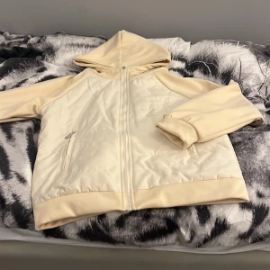 Beige quiltad jacka med huva - Säljer en stilren beige jacka med huva och dragkedja framtill. Jackan är quiltad och har långa ärmar samt ribbade muddar vid ärmslut och nederkant. Perfekt för kyligare dagar!