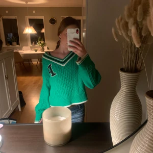Grön stickad tröja - Snygg grön stickad tröja med en stor bokstav 'P' på bröstet. Tröjan har en v-ringad krage med vita och svarta ränder och är croppad i modellen. Perfekt för en avslappnad stil.