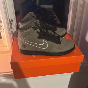 Nike Dunk - Säljer ett par Nike Dunk i grå mocka med svarta detaljer och vit swoosh. Skorna har snörning och en unik design med tryck på hälen. Perfekta för dig som vill ha en stilren och modern look. Använda en gång, digitalt kvitto finns.