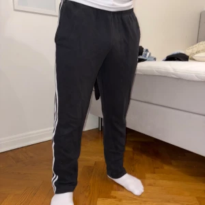 Svarta mjukisbyxor från Adidas - Säljer ett par svarta mjukisbyxor från Adidas med de klassiska tre vita ränderna längs sidorna. Byxorna har en elastisk midja och är perfekta för en avslappnad stil. De är tillverkade i ett mjukt material som ger komfort hela dagen.