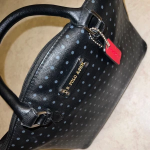 Svart prickig handväska från U.S. Polo Assn. - Vintage svart handväska med prickigt mönster från U.S. Polo Assn. Väskan har en dragkedja och ett rött foder inuti. Perfekt för att ge en stilren touch till din outfit.