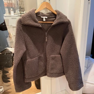 Brun teddyjacka med dragkedja - Mysig brun teddyjacka med dragkedja och stora fickor framtill. Perfekt för kyliga dagar när du vill hålla dig varm och bekväm. Jackan har en hög krage och långa ärmar för extra värme.