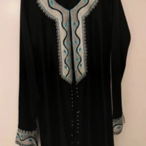 Svart klänning med broderade detaljer (Abaya) - Elegant svart klänning med vackra broderade mönster i turkos och silver längs halsringningen och ärmsluten. Klänningen har långa ärmar och en unik design med fransar framtill. Perfekt för speciella tillfällen.