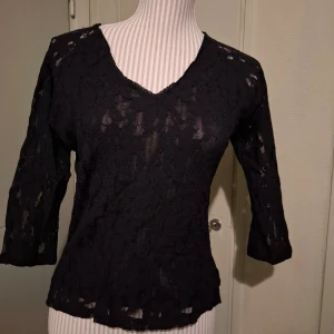 Svart spetsblus  - Snygg svart spetsblus från Pepper corn med v-ringning och trekvartsärmar. Perfekt för en stilren look med en touch av elegans. Blusen har ett genombrutet mönster som ger en fin detalj.