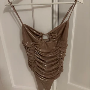 Topp Zara - Säljer en snygg beige glansig body från Zara med smala axelband och rynkade detaljer. Toppen har en liten cut-out framtill och är perfekt till fest! 